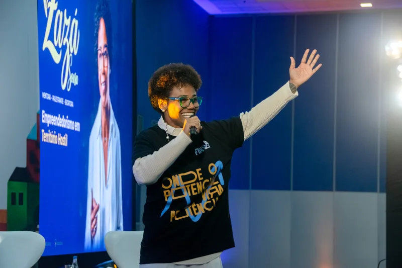 Zazá Sousa sorrindo no palco com telão ao lado — Favela Mais Rica